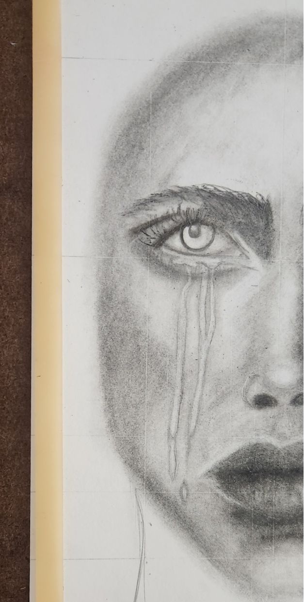 Early stage of Behind the Mask. Half of a face is shaded in graphite over a pencil grid, with tears running from an eye that has not yet been coloured.