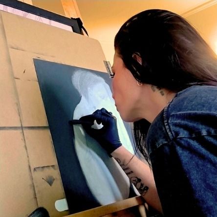 Melissa at an easel, drawing a figure in white on dark paper, wearing black gloves.