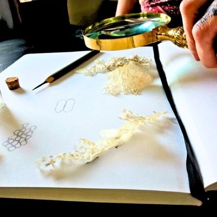 An open sketchbook with pencil studies and strips of lace, beside a brass magnifying glass.